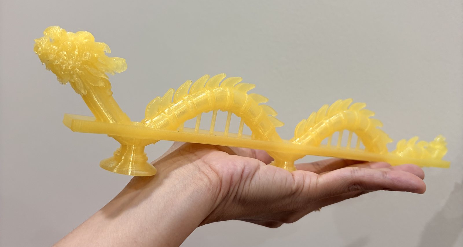 A whole yellow translucent printed dragon bridge lying across an open hand: the frilly head on the left, two ridged arches over a flat deck with thin hangers, three round feet and a flame-shaped tail
