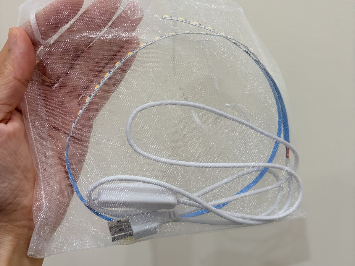 A hand holding a sheer white bag with a coiled LED strip inside: a thin strip with tiny yellow lights on a blue backing, a white cable, an inline switch and a USB plug