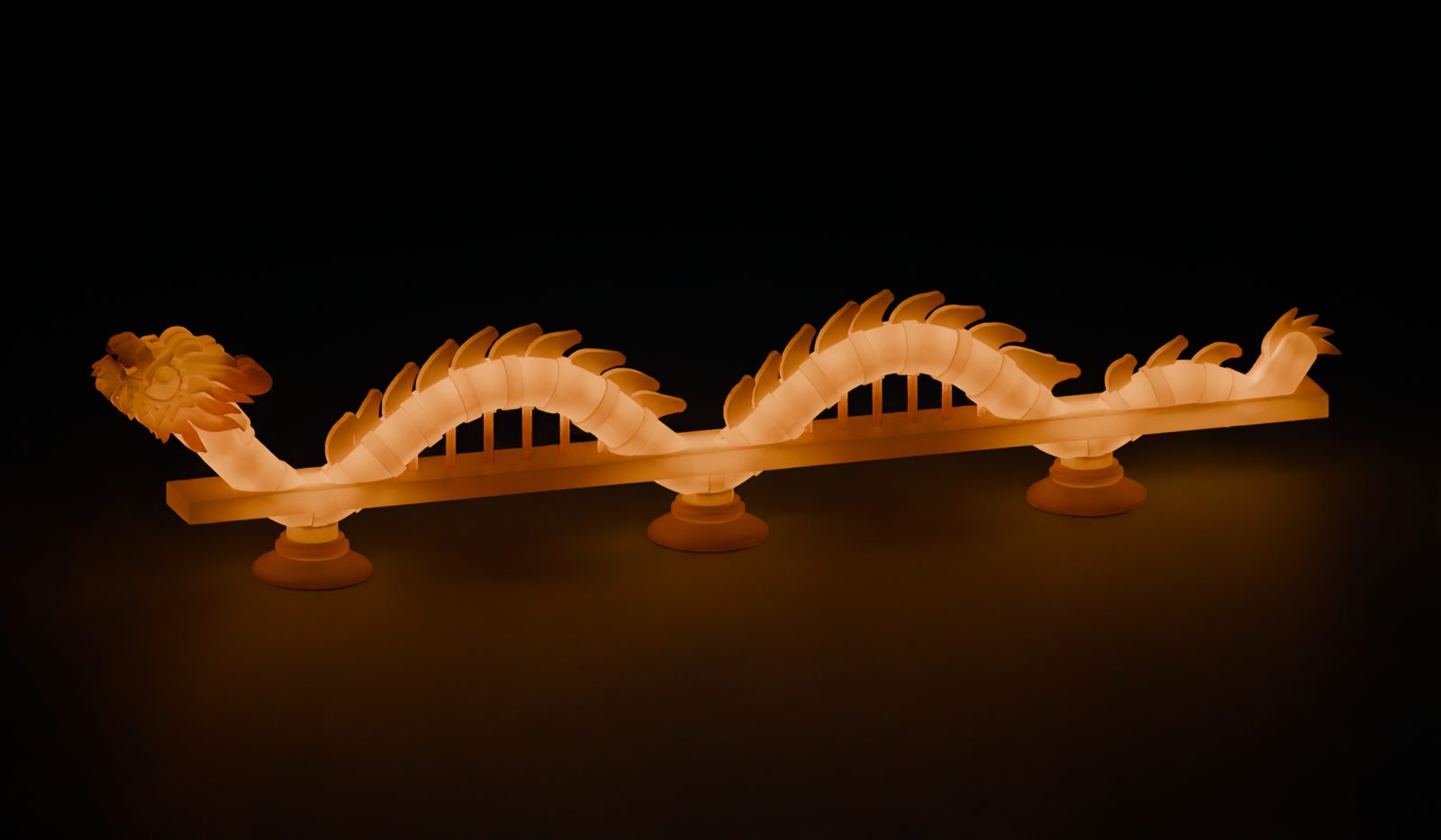 A render of the lamp at night: a golden dragon with a spiky mane and a ridged back, glowing orange from inside, curving over a flat bridge deck on three round feet