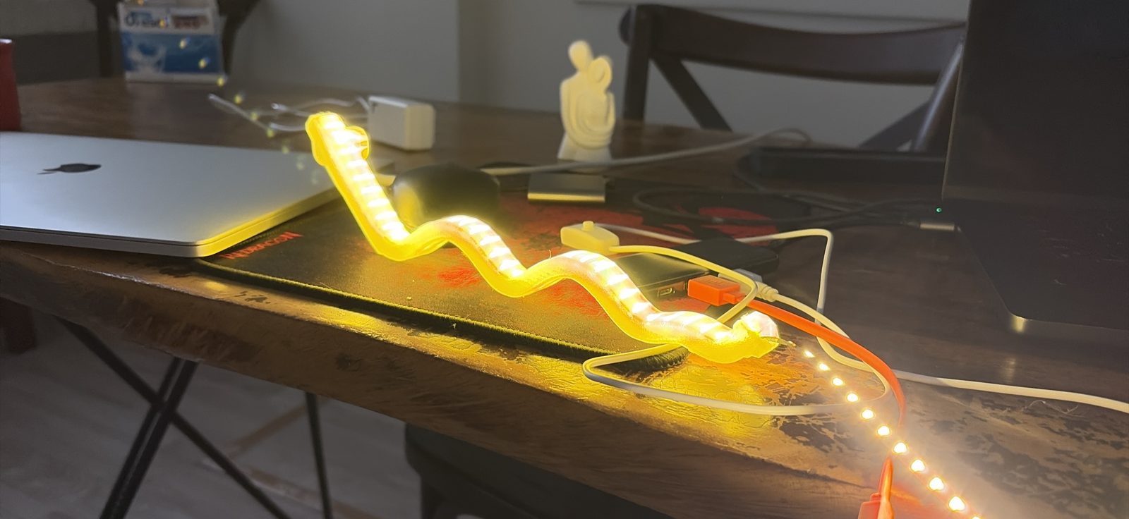 On a wooden desk next to a closed laptop, a wavy translucent yellow tube glows along its whole length, with the LED strip's lit end and white USB cable coming out of it