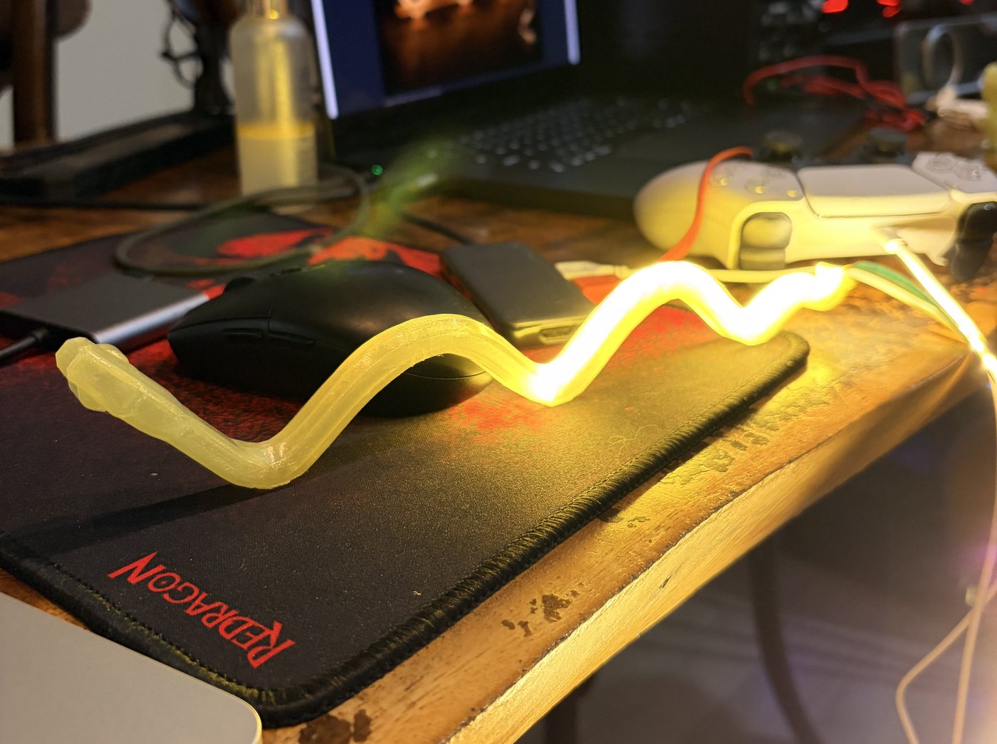 On a desk with a mouse pad, a wavy yellow test tube lies with the LED strip inside: the right half glows brightly, the left half stays dark where the strip never reached