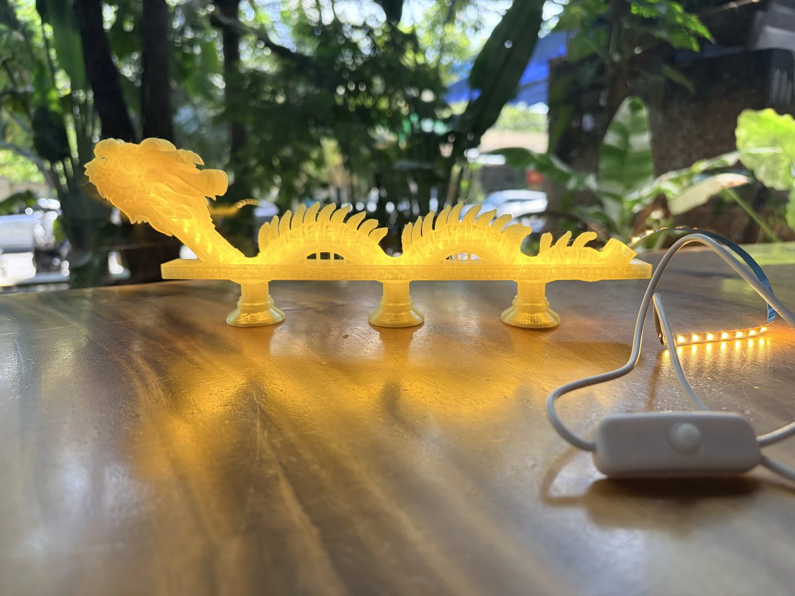 Chiếc đèn đang sáng trên bàn cà phê ngoài trời, phía sau là cây xanh, bên cạnh là dải đèn và công tắc