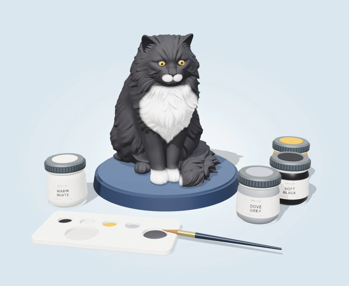 The finished cat figurine, black and white with amber eyes, beside four pots of paint, a palette and a brush