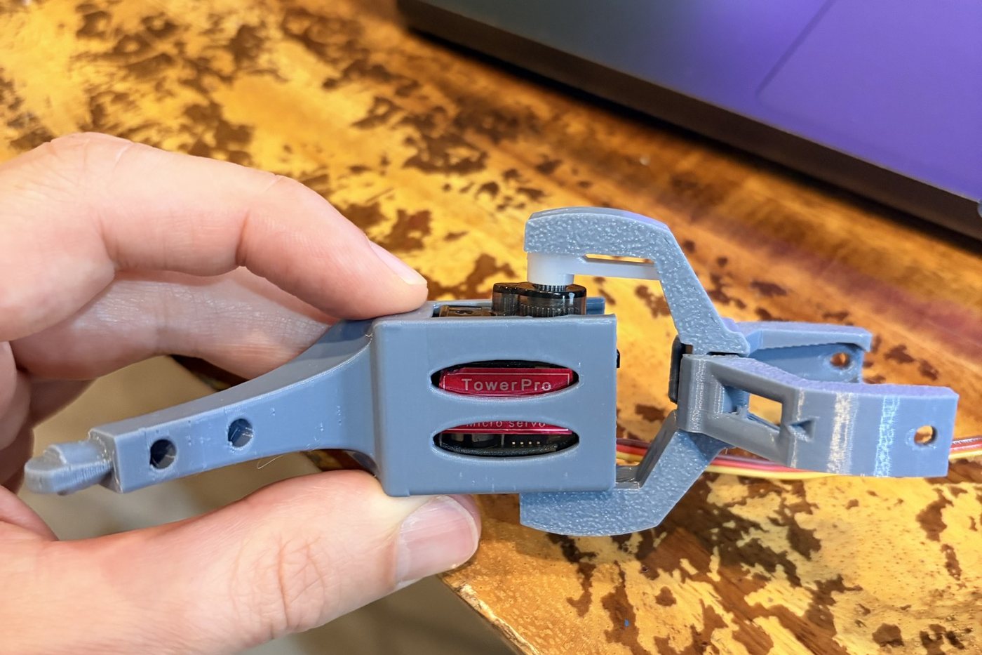 A blue 3D-printed robot leg with a small red-labelled servo motor inside, held in a hand over a wooden table