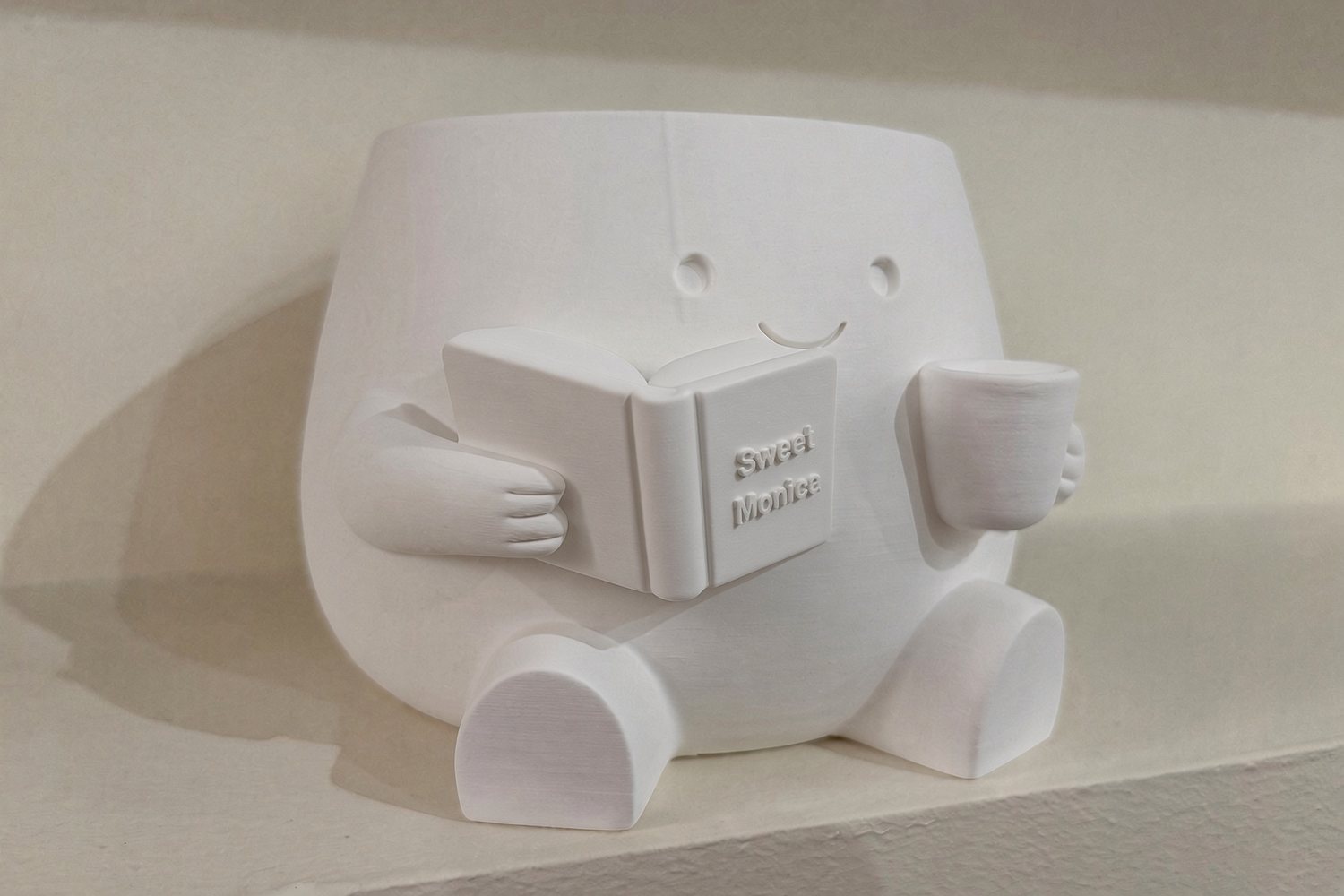 A white printed planter shaped like a smiling reader holding a mug and a book that says Sweet Monica